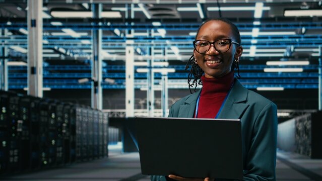 Portrait of happy data center system administrator doing resource optimization using laptop software, Smiling young employee in server hub using notebook to upgrade IT infrastructure, camera A.