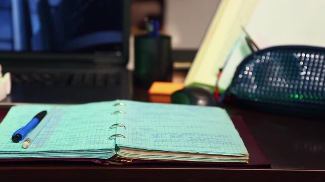 An open notebook with pen on the school desk with a laptop, headphones and work or study supplies. No people, nobody. A detail of the workspace on a wooden table. Close-up motion atmosphere video