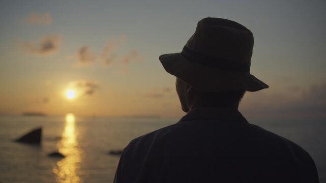Silhouette of a man wearing a hat while watching the sunset over the ocean horizon, capturing calm evening reflections and a peaceful tropical travel atmosphere.
