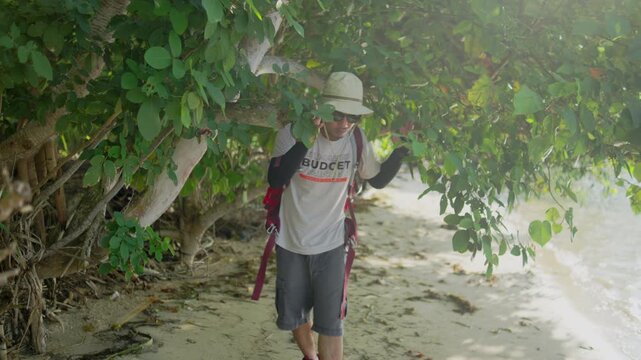A backpacker exploring a tropical beach beneath dense green foliage, wearing a hat, sunglasses, and backpack while enjoying a sunny outdoor travel adventure.