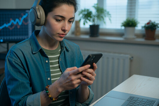 Young woman wearing headphones using smartphone beside laptop, managing mobile workflow at home office. Ideal for remote work, digital communication, productivity content