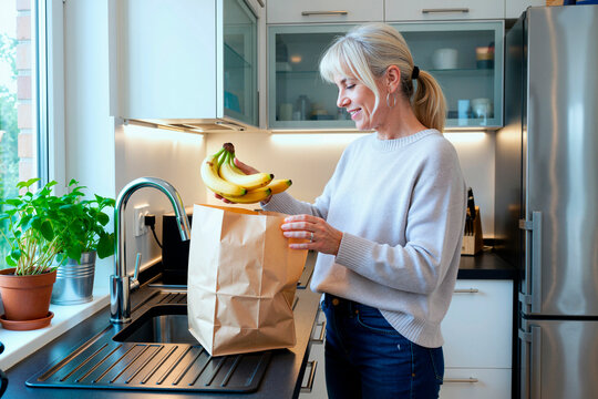 Adult woman unpacking bananas from grocery paper bag in kitchen, supporting healthy eating and home food shopping. Useful for nutrition, retail, lifestyle marketing