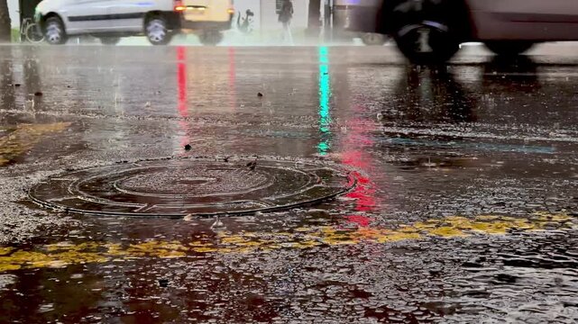 Flooded city street at night with a manhole cover surrounded by rainwater ripple, car passing in background, rain drop splashing on wet pavement
