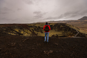 Une voyageuse en veste rouge observe un vaste cratère volcanique depuis son bord. Le sol sombre et les reliefs volcaniques contrastent avec le ciel gris chargé de nuages. © Tof - Photographie