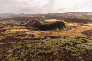Vue panoramique d’un ancien cratère volcanique entouré de champs de lave recouverts de mousse. Les reliefs volcaniques aux teintes noires, rouges et ocres contrastent avec les vastes plaines islandais © Tof - Photographie