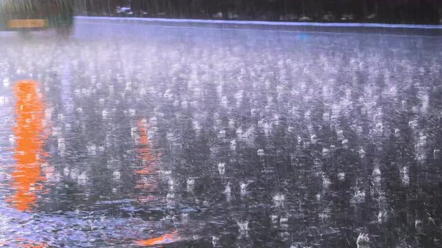 Close view of wet asphalt road surface during heavy rain with colorful orange and red light reflection, rain drop splash pattern and passing car wheel visible