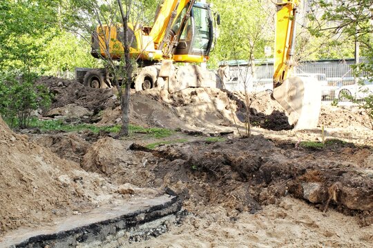 A yellow wheeled excavator operating on a city construction site with piles of dug soil, sand, broken asphalt layers and a metallic bucket in the foreground surrounded by green deciduous trees