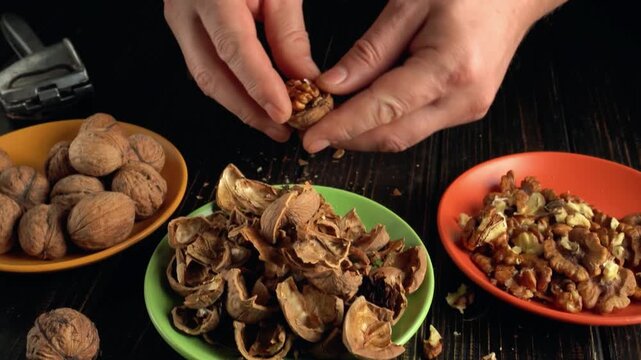 Hands of a person cracking open walnuts, separating shells and nuts into colorful bowls on a dark wooden table