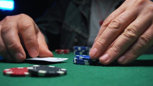 Hands of a male poker player placing chips on a green felt table while holding playing cards in a dimly lit casino environment