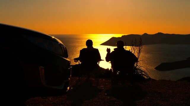 Silhouette of friends watching sunset over sea on a road trip. Two silhouetted friends relaxing in camping chairs next to their car on a cliff, enjoying the stunning golden sunset over the ocean