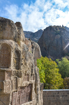 Intricately carved khachkars are etched into the rock walls of Geghard Monastery. Towering cliffs and lush green trees rise in the background under a soft, cloudy sky, highlighting ancient artistry