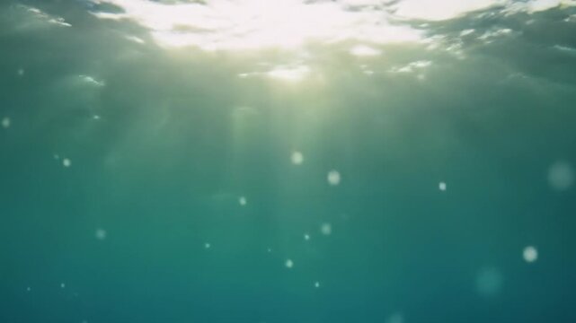Low-angle wide underwater video looking up at the ocean surface. Radiant golden sunbeams pierce deep blue-green water, creating sharp light rays with drifting luminescent particles. Very slow push-in