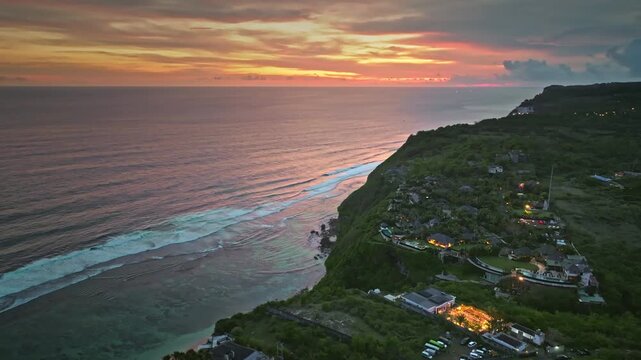 Bali, Bukit Peninsula coastline with luxury cliffside villas and resorts glowing at sunset, lush green cliffs meeting the Indian Oceans rolling waves under vivid sky. Aerial view drone flight panorama