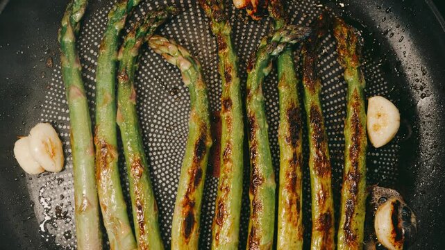 Asparagus frying in a pan with garlic cloves and visible steam. Charred edges and sizzling oil create a rustic cooking scene with rich texture.