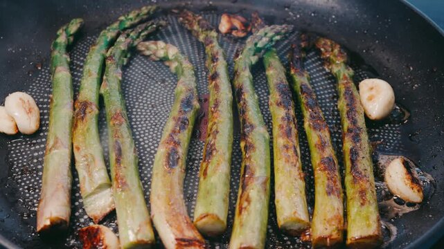 Asparagus frying in a pan with garlic cloves and visible steam. Charred edges and sizzling oil create a rustic cooking scene with rich texture.
