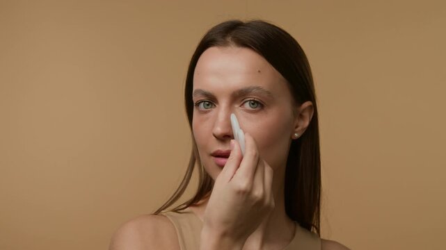 Close-up portrait of Caucasian brown-haired gen Z girl looking at camera using white jade stone massage tool against beige background