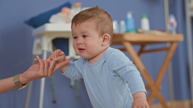 Boy holding an adult's hand and reaching forward while learning to walk beside a changing table and baby bottles in a nursery; determination trust.