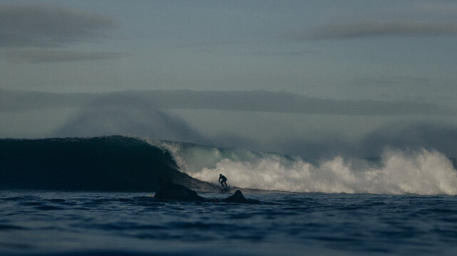 surfer paddling by as another surfer rides a large wave at sunrise