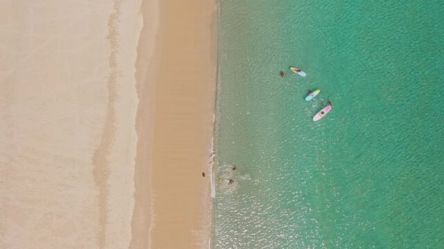 People are swimming, paddleboarding, and relaxing in the clear turquoise water and golden sand of Karon Beach, Phuket, enjoying a sunny day during tropical holidays. Aerial top down view drone flight