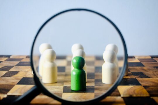 High-angle shot looking through a magnifying glass at a green wooden pawn surrounded by blurred white figures on a chess board.