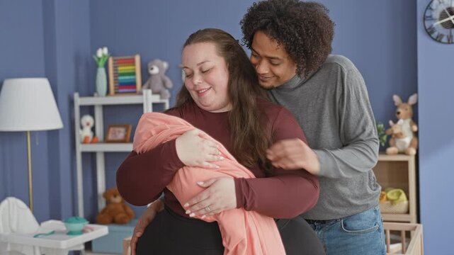 Plus size woman cradles swaddled baby while father embraces from behind in nursery with toys and shelf visible; family love.