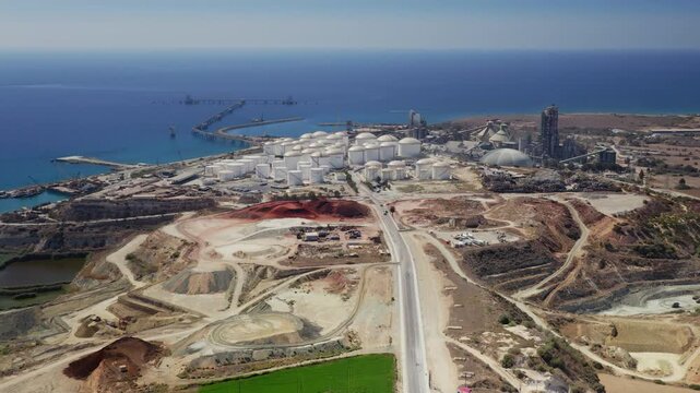 Aerial view drone flight over large industrial complex along the coast of Cyprus, featuring numerous fuel storage tanks, a prominent cement factory, and active quarry operations under a clear blue sky