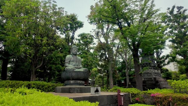 A serene bronze Buddha statue sitting on a lotus pedestal surrounded by lush green trees in a Japanese temple (Japanese text on the small wooden sign is unreadable)