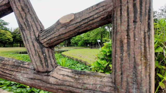 Beautiful park landscape with a pond and fresh greenery viewed through the gap of a rustic wooden-style railing with foreground blur