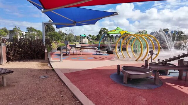 Children's splash park featuring active water jets spraying, beneath tensile shade sails in a suburban neighborhood of Melbourne Australia. Family-friendly recreational equipment and community space