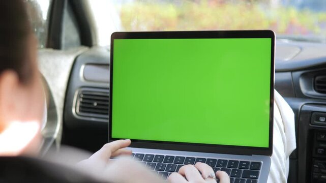 Person uses laptop with green screen inside car. Hands type on keyboard, focused on work. Ideal for remote work, travel, or digital presentations. Green screen allows easy video editing