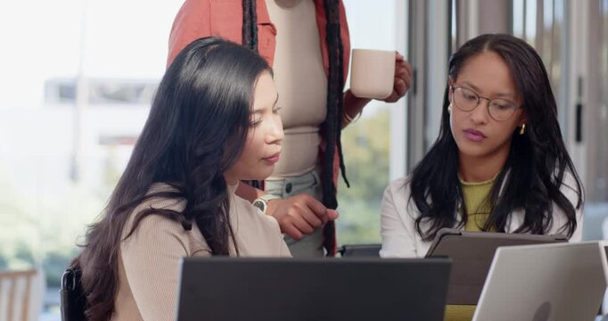 Diverse women at laptops in office shifting, colleague coming with mug pointing tablet explaining