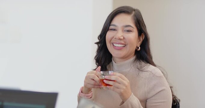 Adult woman holding glass teacup, smiling and reacting to off-camera speaker in office with laptop