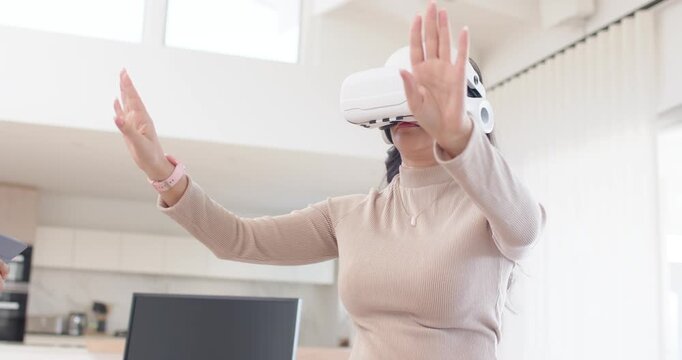 Diverse female team watching tablet, guiding headset user in VR doing gestures in bright kitchen