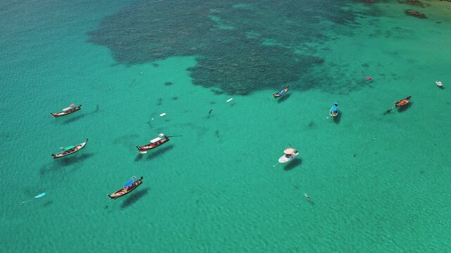 Traditional longtail boats and yacht floating on the clear turquoise tropical sea over visible coral reef, with people swimming and enjoying the vacation in Phuket, Thailand. Aerial view drone flight