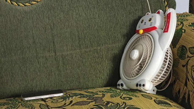 White cat shaped electric fan on patterned sofa next to smartphone