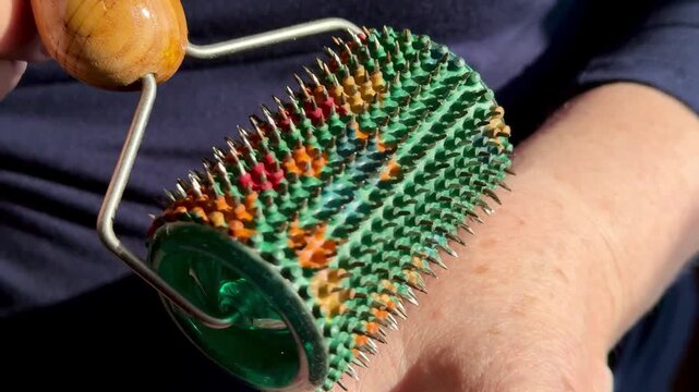 Close-up woman hands rolling green needle roller, wooden handle guiding colorful spikes across skin, slow rhythmic motion