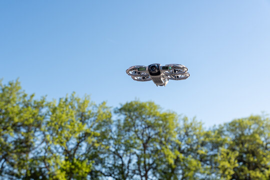 A drone with protective circular guards around its propellers hovering against a clear blue sky, featuring a central camera for aerial footage, with leafy green trees below indicating a natural spring