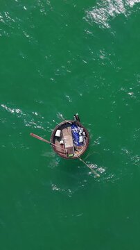 Vertical screen Aerial View Hoi An Coracle On Emerald Sea, Lone Fisherman Rows With Wooden Paddle, Tethered Net Trailing, Sunlight Glittering On Green Water, Traditional Round Craft Floating