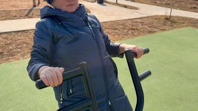 Elderly Caucasian woman exercising at stadium on outdoor leg and arm machine during spring day, bundled in warm coat, hat