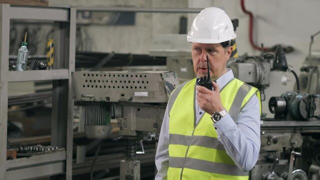 Industrial supervisor in safety helmet using walkie talkie inside manufacturing workshop with machinery in background.