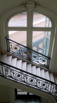 Interior view of an elegant Parisian staircase with ornate iron railings and a large arched window bringing soft daylight into the building.