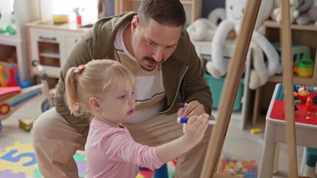 Man guiding girl holding marker at easel in a colorful playroom studio, pointing pen while teaching drawing; learning concentration.