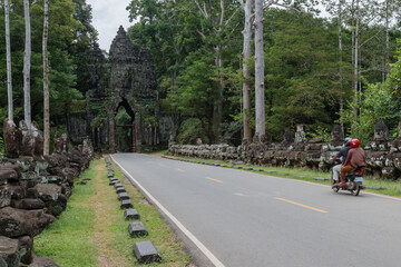 Obraz premium Two people ride a motorcycle past the ancient stone statues lining the road to the Angkor Thom North Gate, Siem Rep, Cambodia.