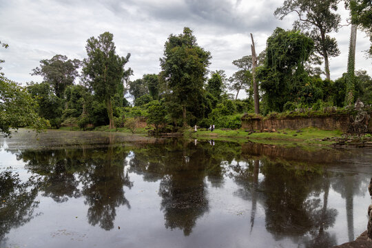 A group of tourists explore the ancient ruins of Preah Khan Temple, Siem Rep, Cambodia. The temple is surrounded by lush jungle and a reflective moat.