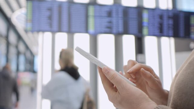 Hands hold smartphone at airport terminal. Background shows flight information screens. Person focuses on device, unaware of surroundings. Traveler waits for departure, checks updates