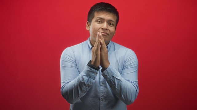 Young hispanic man with clasped hands in a pleading gesture against a red studio backdrop; hopeful apology.