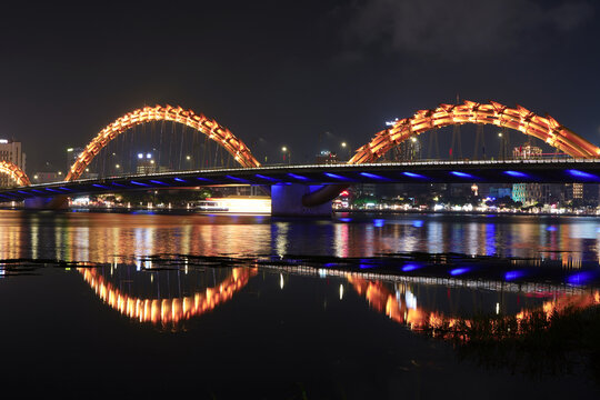 A massive illuminated Dragon bridge spans across a wide river at night in Da Nang, Vietnam, creating a vivid reflection on the water