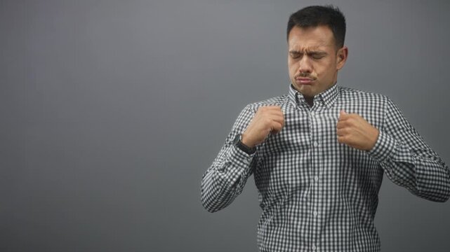 Hispanic man sniffs checkered shirt, pulling collar with hands at nose in studio; disgust hygiene discomfort.