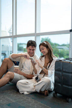 Asian woman girlfriend smiling at phone with white man boyfriend an luggage sits on floor in airport