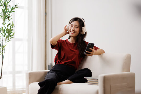 Young asian woman enjoying music, wearing headphones and holding a smartphone, relaxing comfortably on a sofa at home, finding joy and personal time